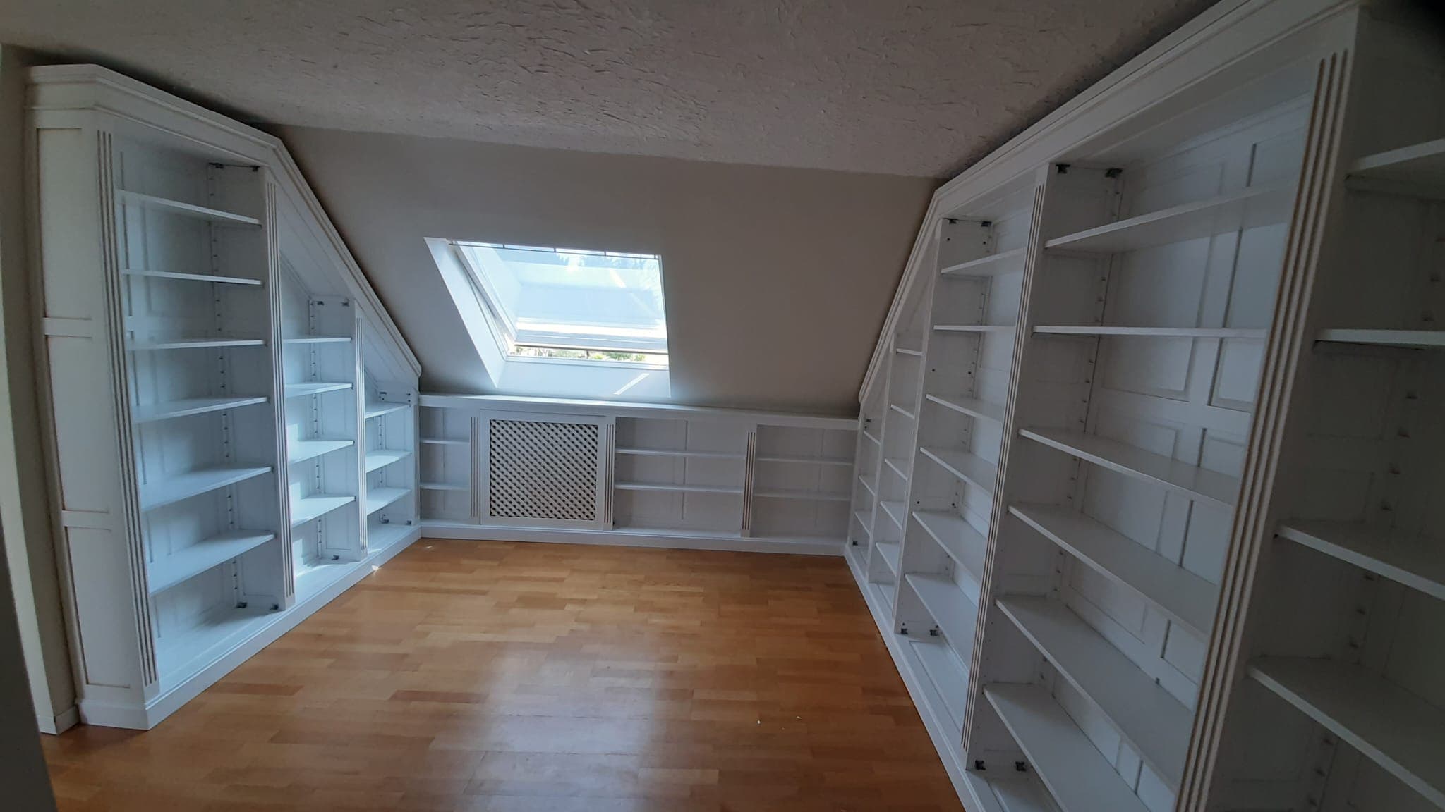 Bibliothèque de comble laquée blanc : corps toute hauteur coupés selon la pente du toit sur les deux murs latéraux, rayonnage bas sous le Velux et cache-radiateur à treillage intégré