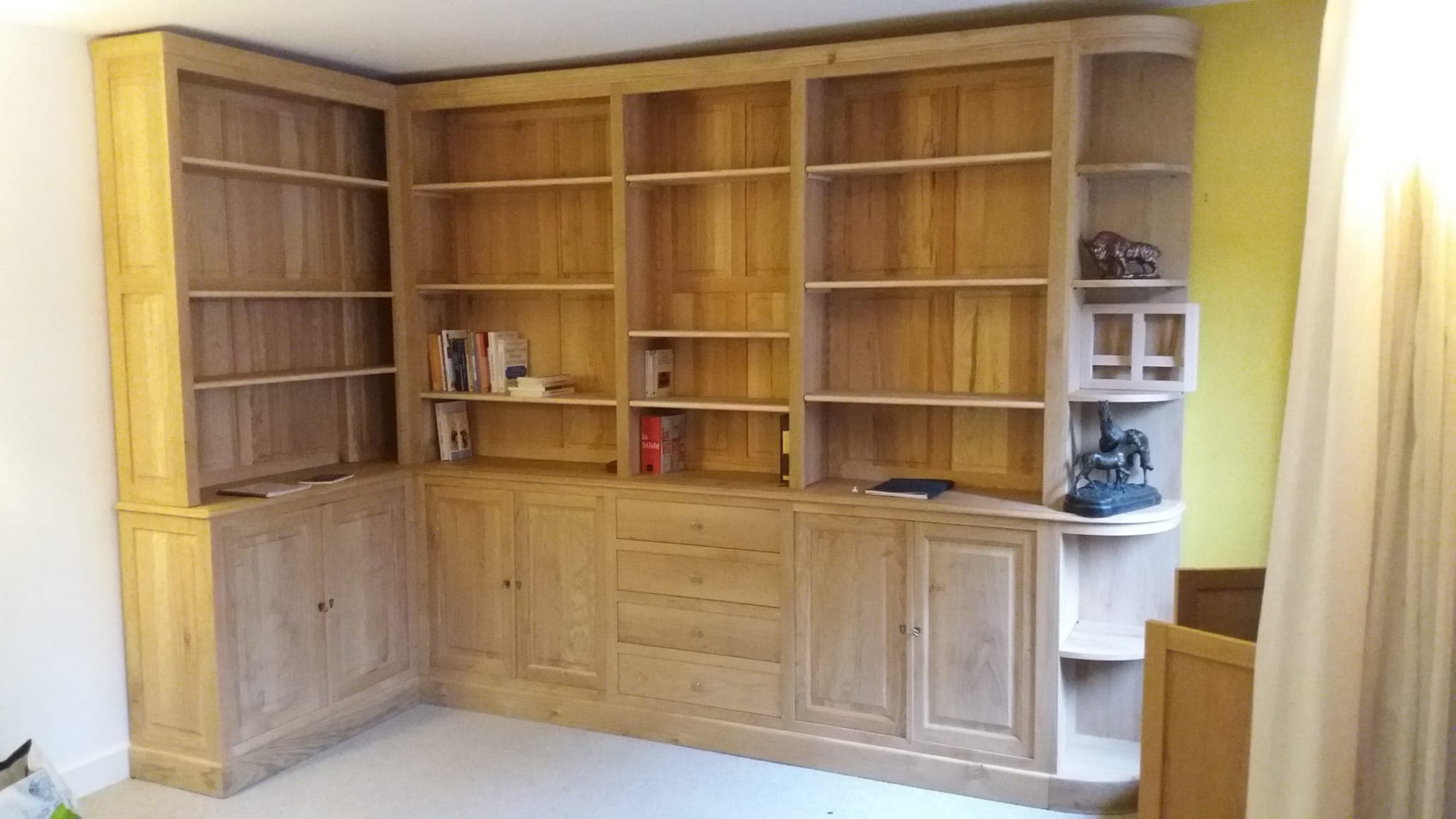 Agencement de bibliothèque d’angle en chêne massif clair : travées de rayonnages à fonds menuisés à panneaux, portes et tiroirs en partie basse, extrémité en retour galbé à tablettes arrondies