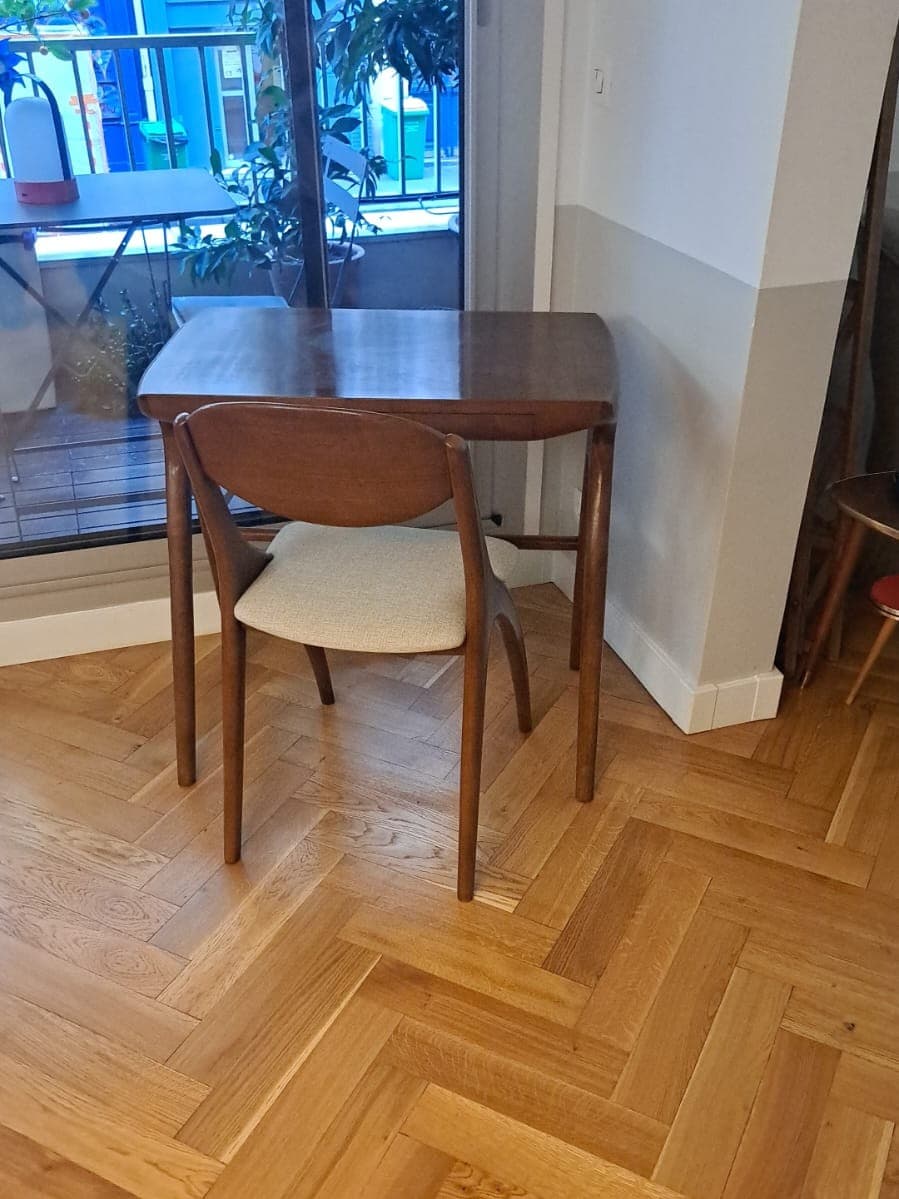 Petit bureau en bois foncé à plateau aux angles arrondis et pieds ronds fuselés, installé devant une porte-fenêtre sur un parquet à bâtons rompus