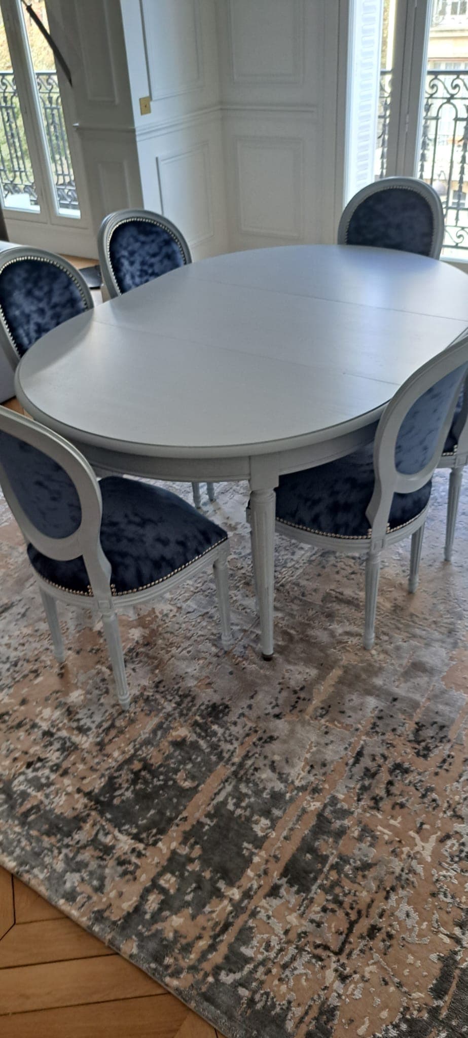 Table ovale Louis XVI laquée gris perle, ouverte sur ses allonges, à pieds fuselés cannelés, entourée de six chaises médaillon garnies de velours bleu nuit