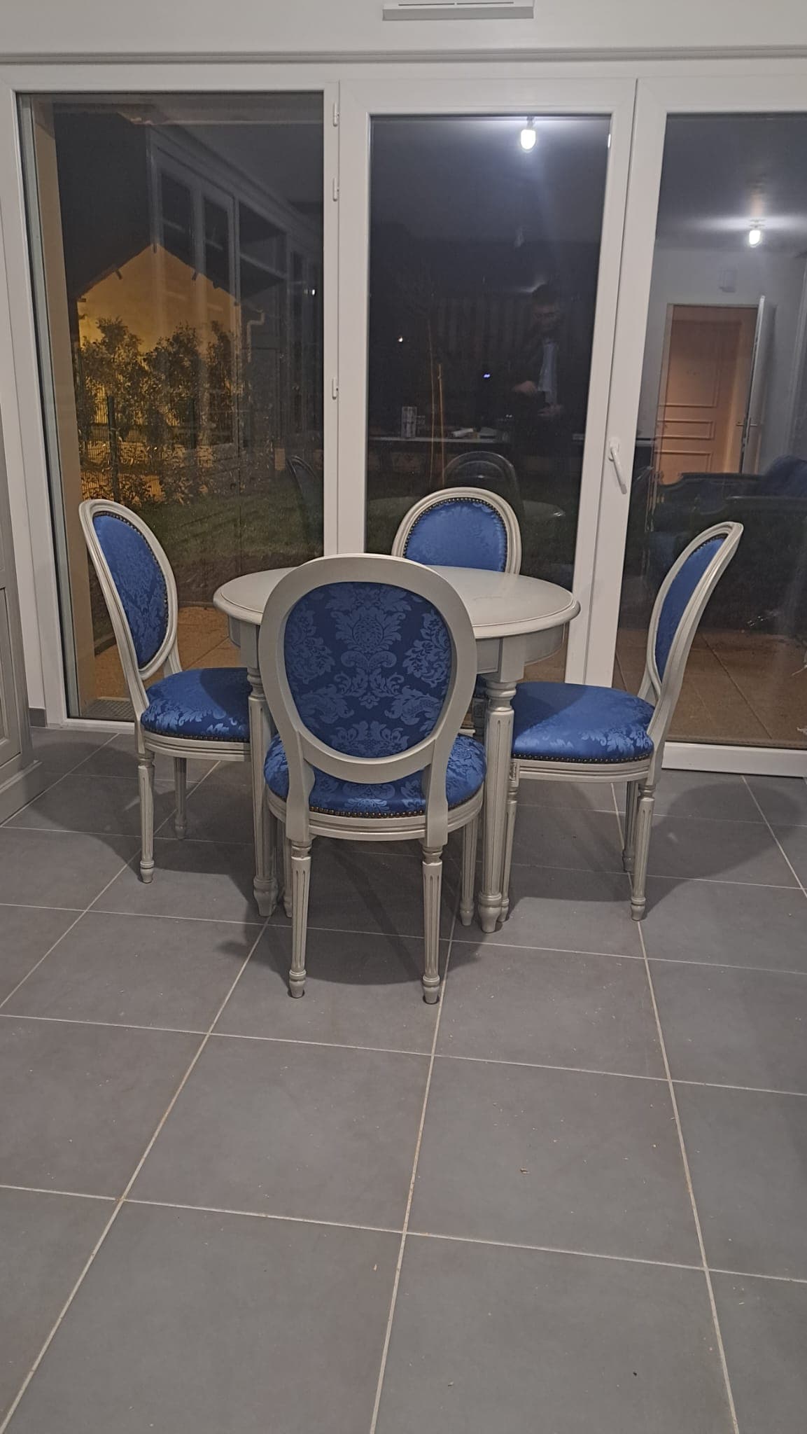 Table ronde Louis XVI laquée gris à pieds fuselés cannelés, entourée de quatre chaises médaillon garnies d’un damas bleu à clous de tapissier